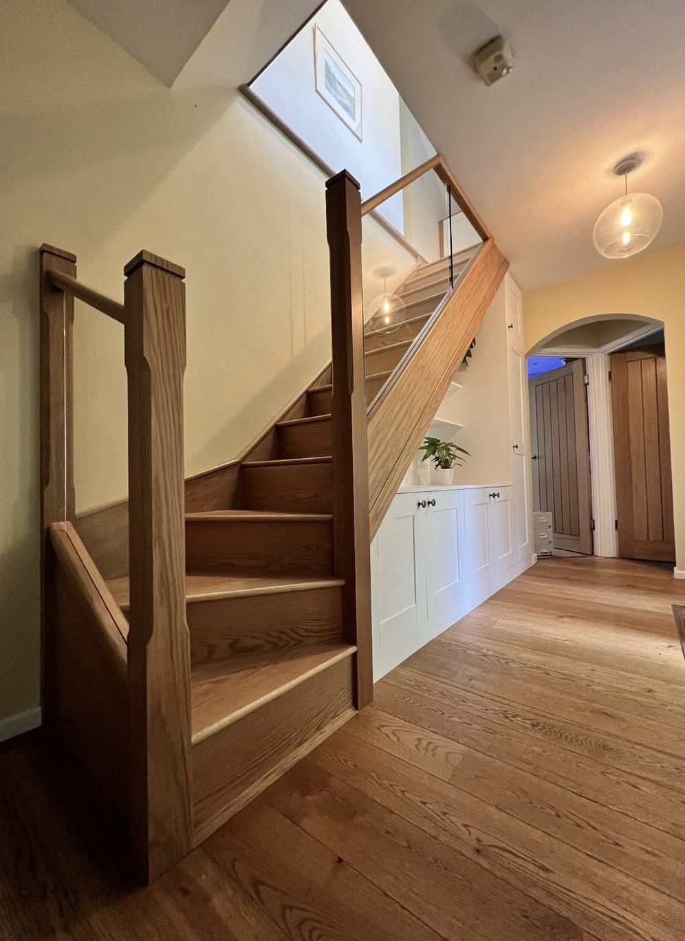 An oak staircase with glass panels rising from a wide timber-floored hallway